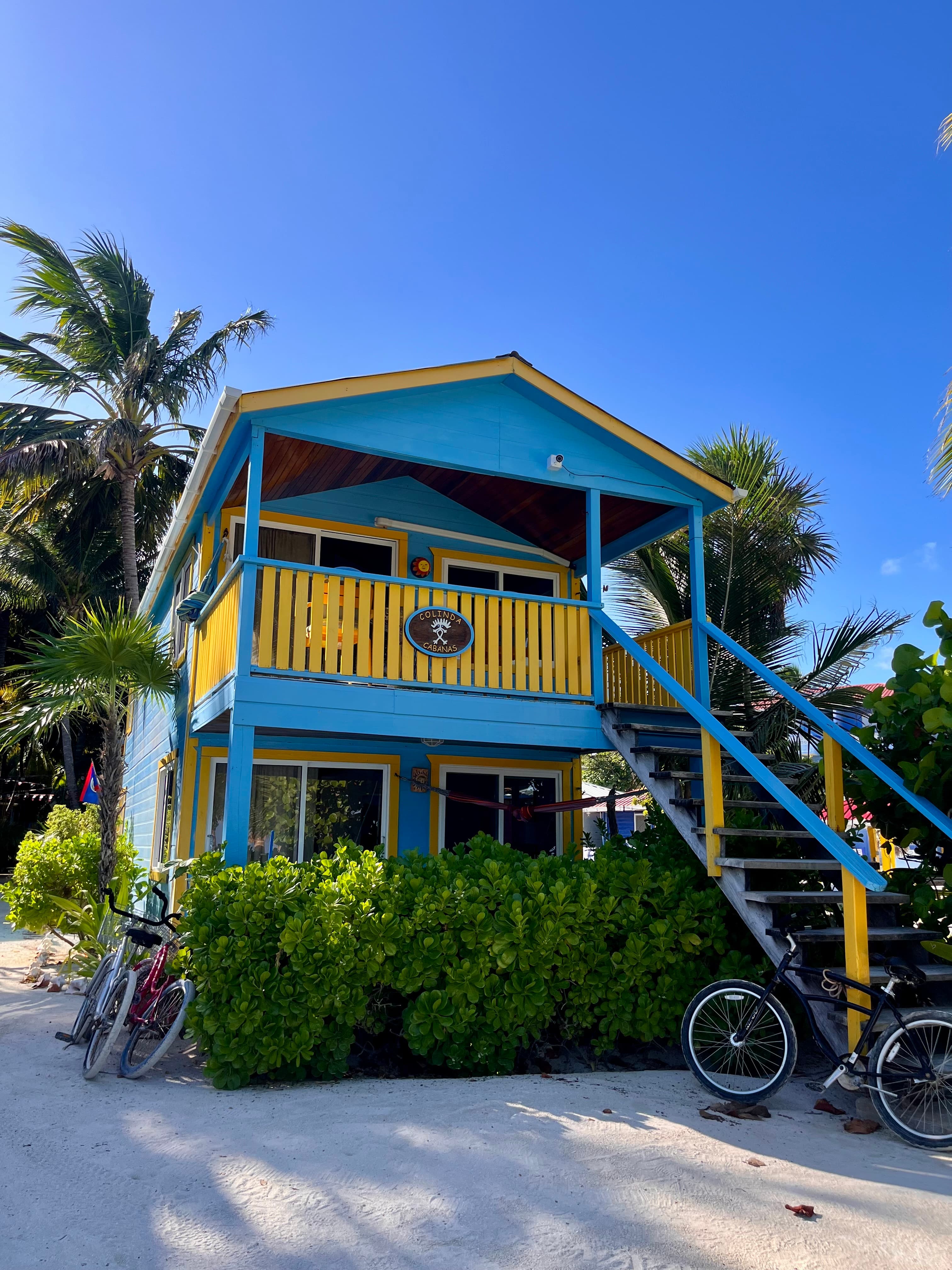 Colorful two-story beach house surrounded by lush greenery and bicycles parked nearby.