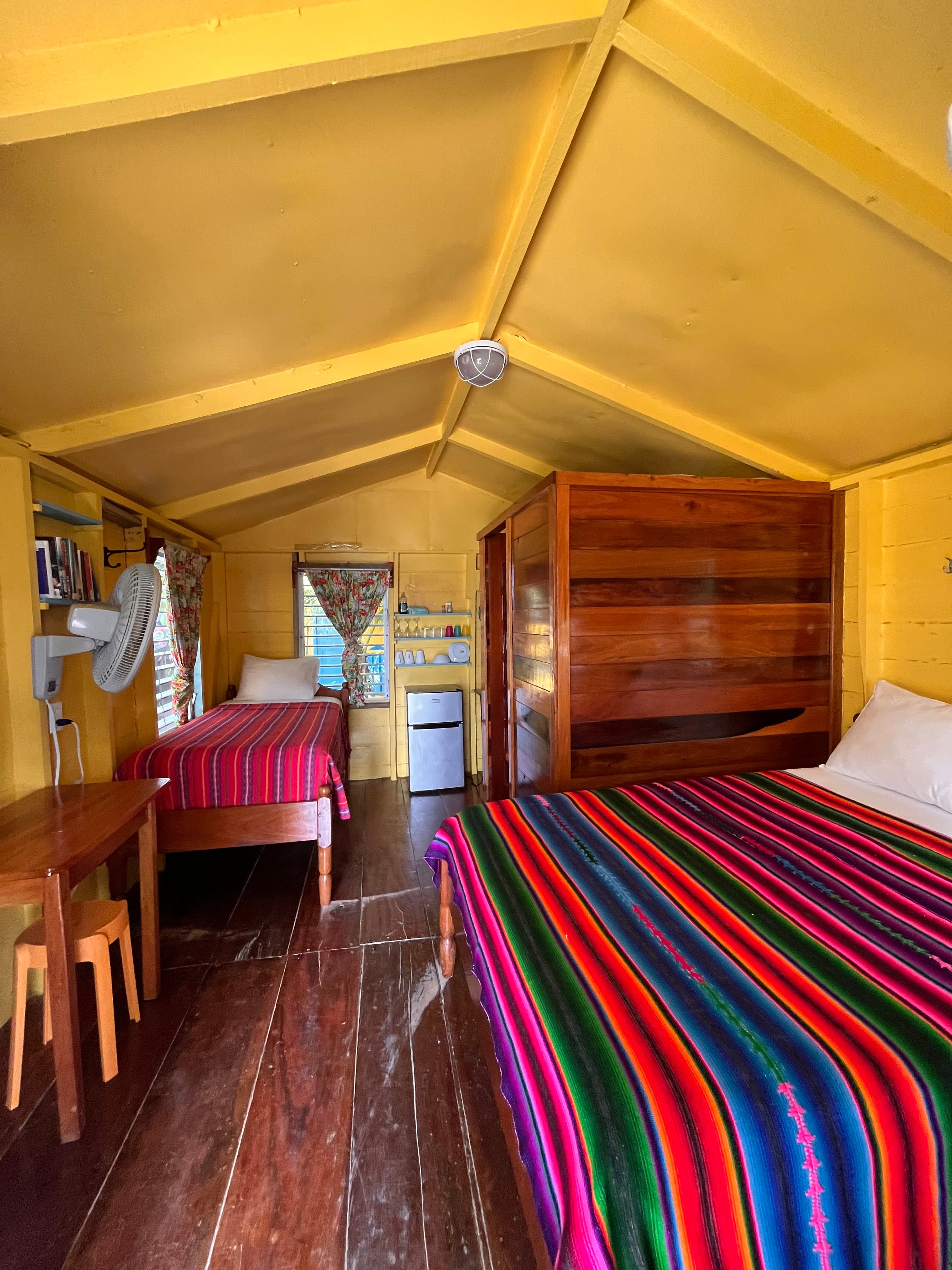 A brightly colored, cozy bedroom with two beds, a wooden table, and a mini-fridge in a yellow cabin.