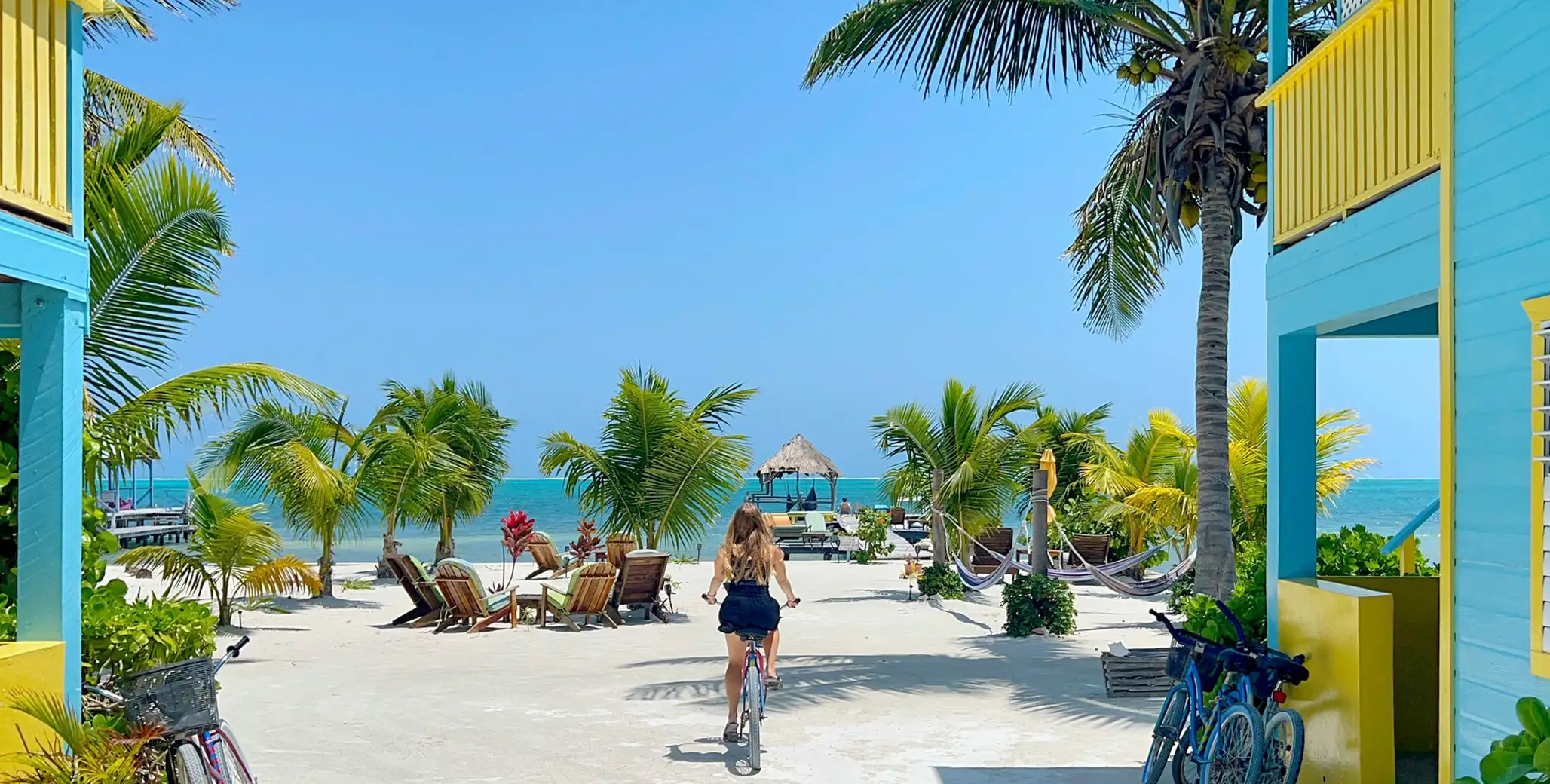 Woman on bike on beach approaching the ocean, hammock and chairs on the beach with teal and yellow building on both sides of the street