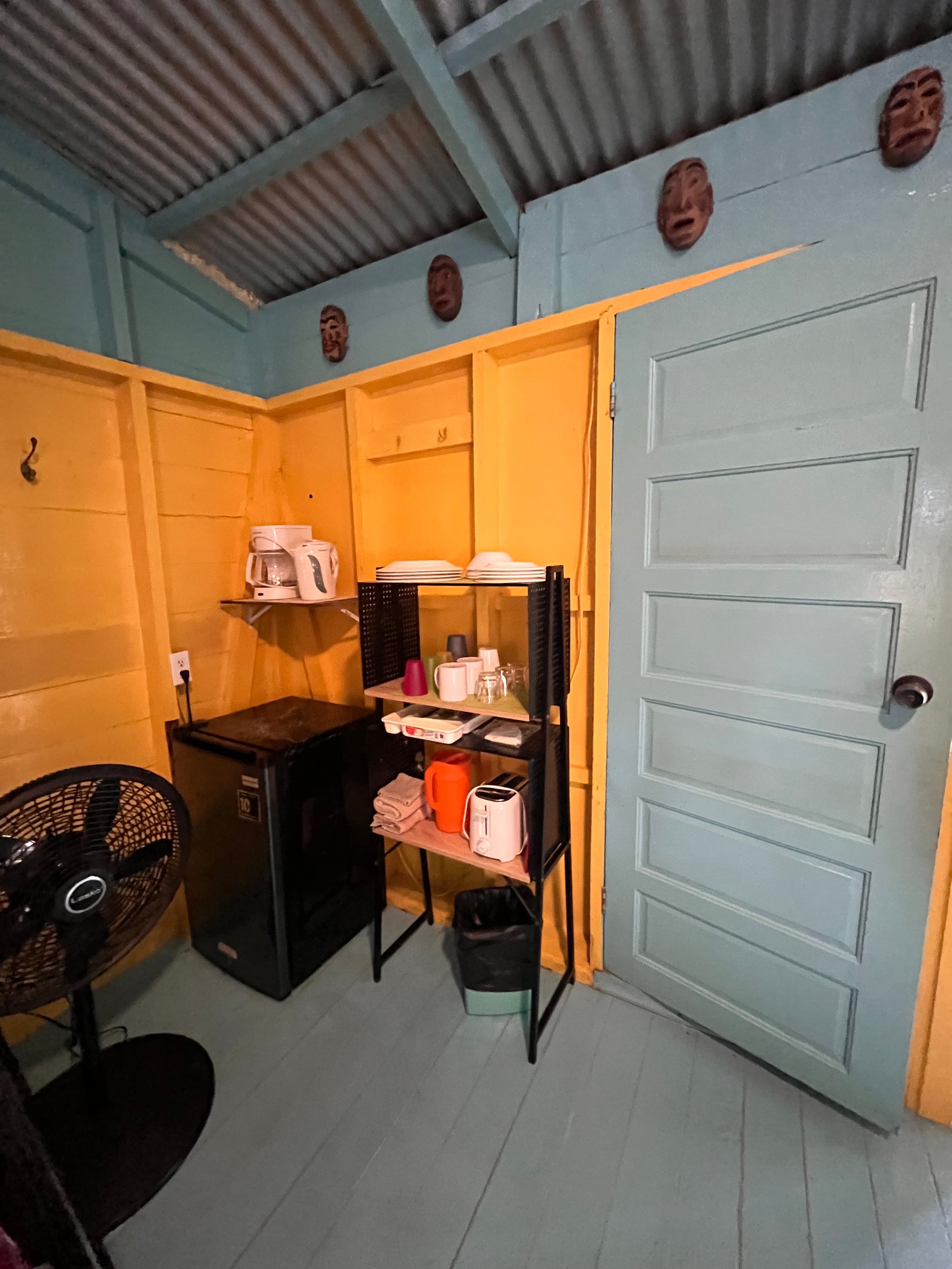 Interior of a cozy room featuring a fan, a mini-fridge, a shelf with kitchen items, and decorative masks on the walls.
