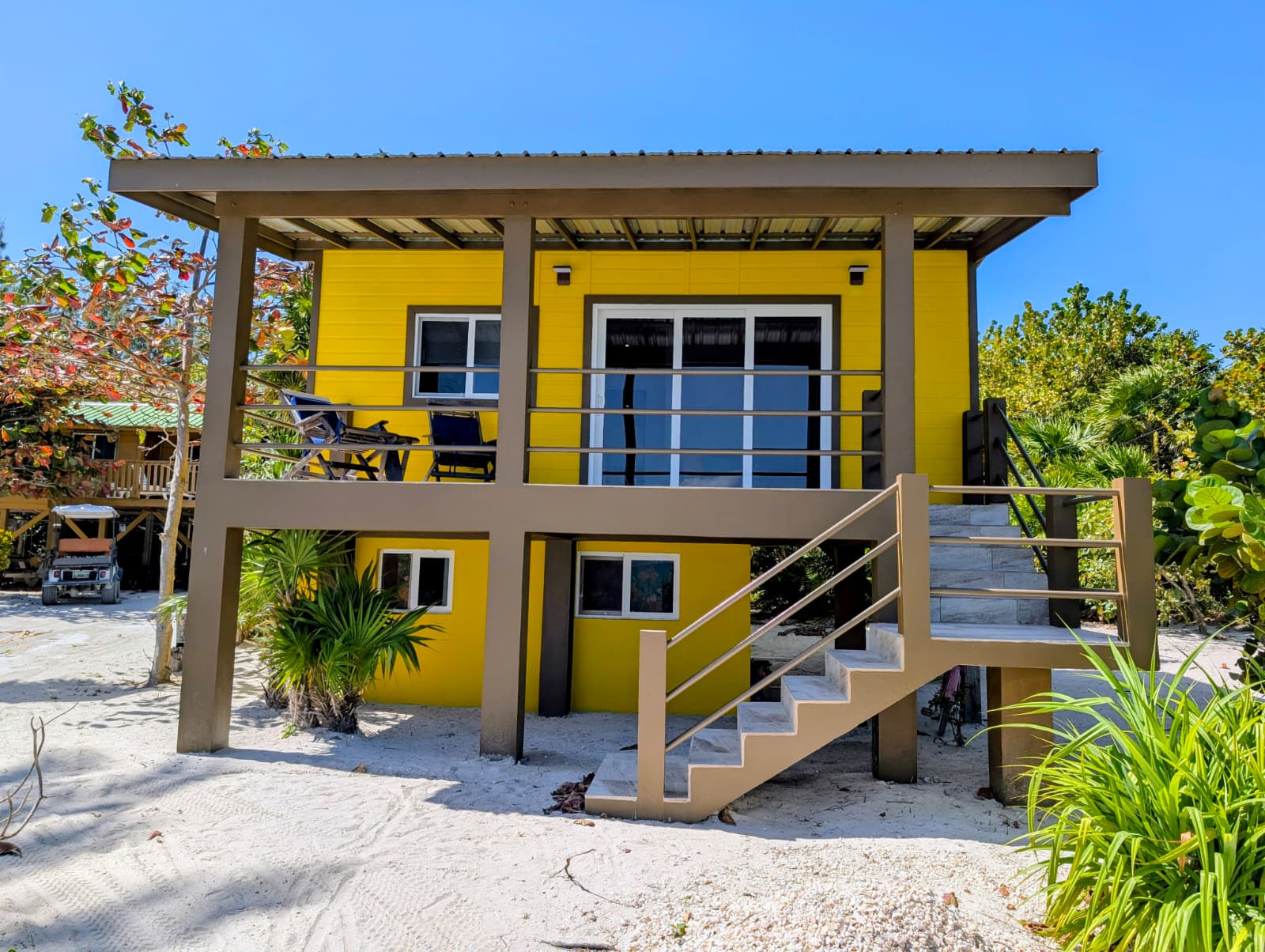 A bright yellow beach house with a staircase and surrounding greenery.