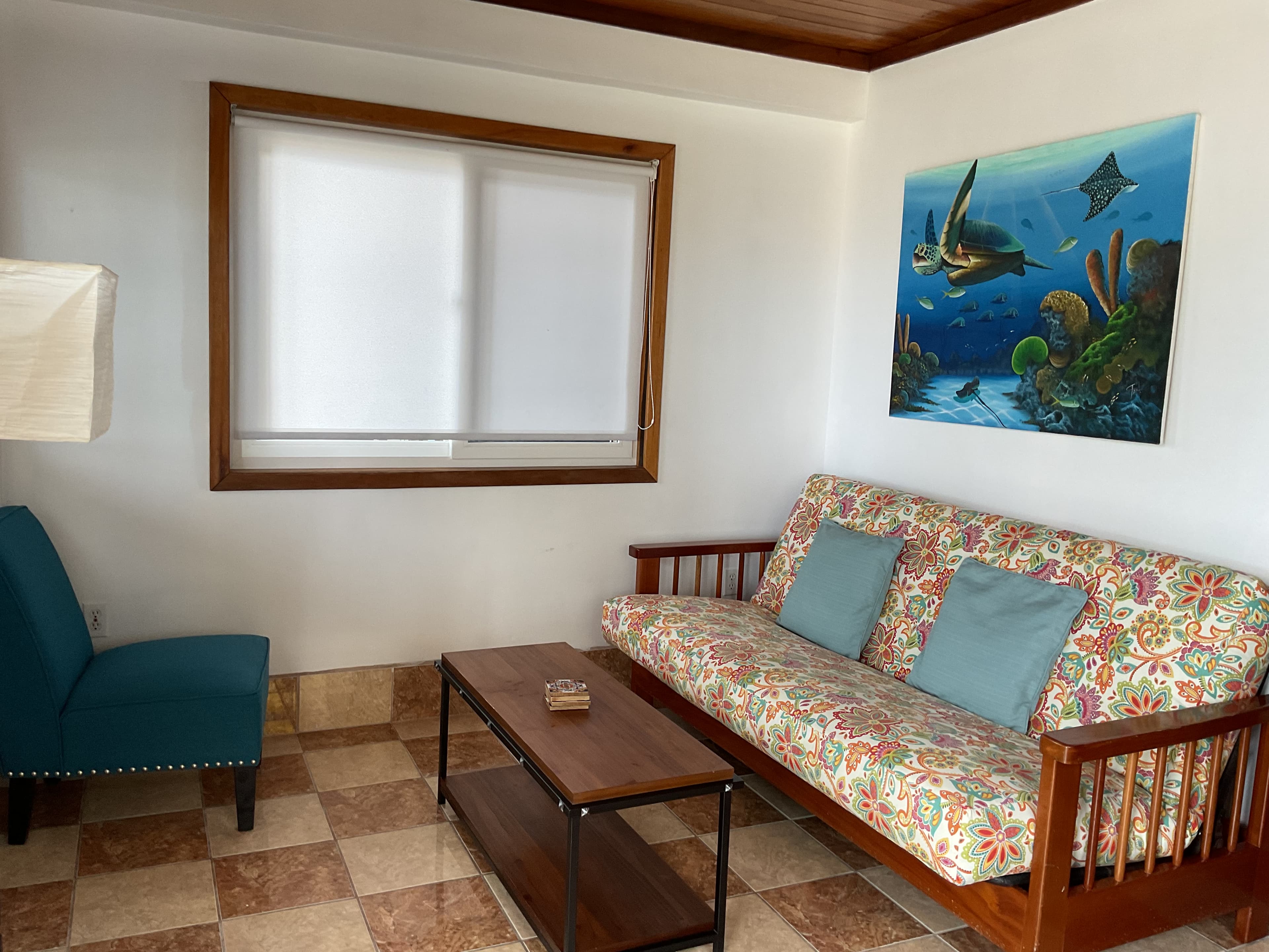 A cozy living area features a floral-patterned sofa, a wooden coffee table, a teal armchair, and an ocean-themed wall painting.