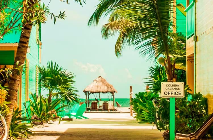A sunny beach path lined with greenery leads to a thatched gazebo over the water, with a sign for "Colinda Cabanas Office" nearby.