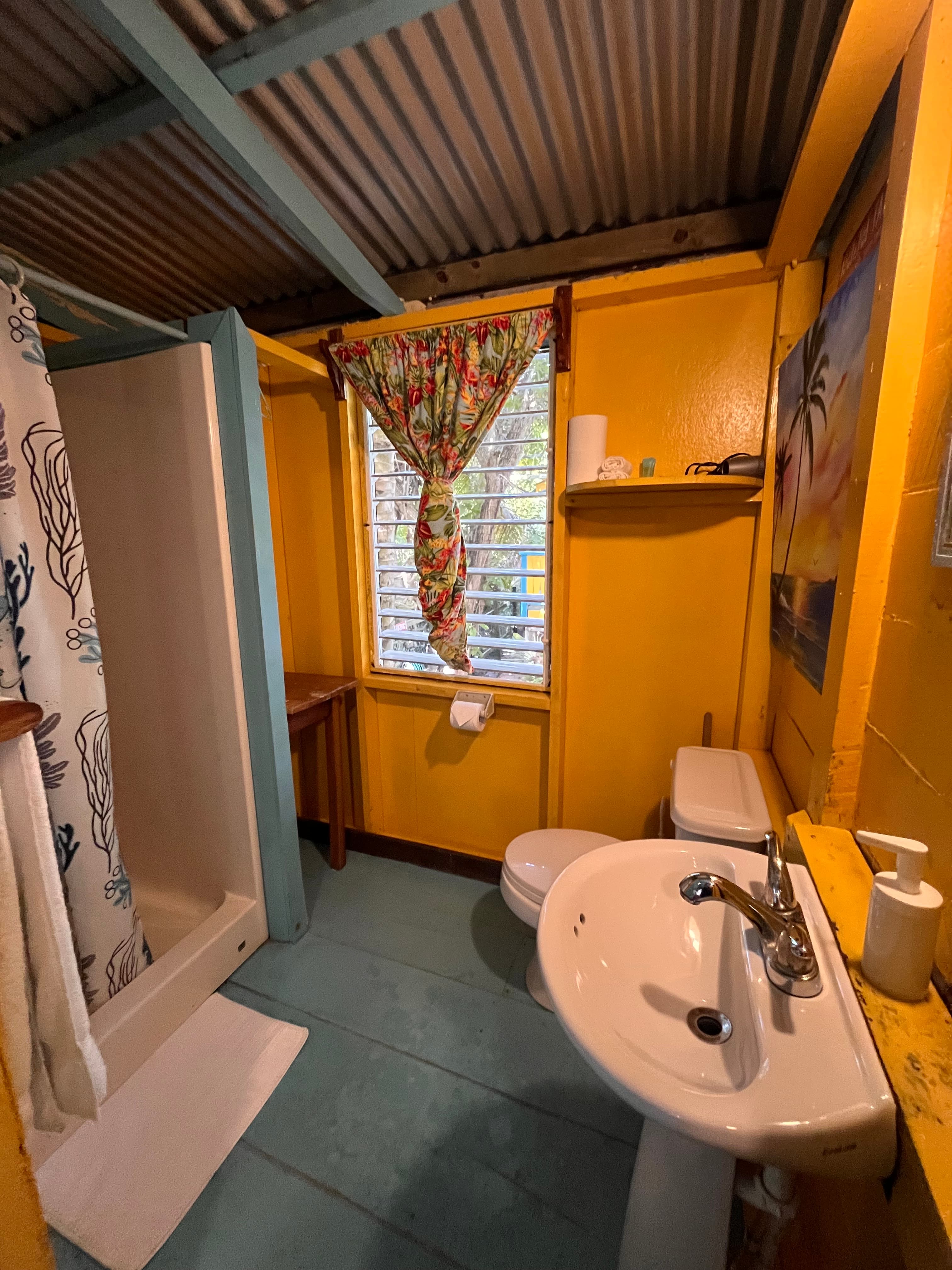 A bright yellow bathroom featuring a shower, toilet, sink, and a window with floral curtains.