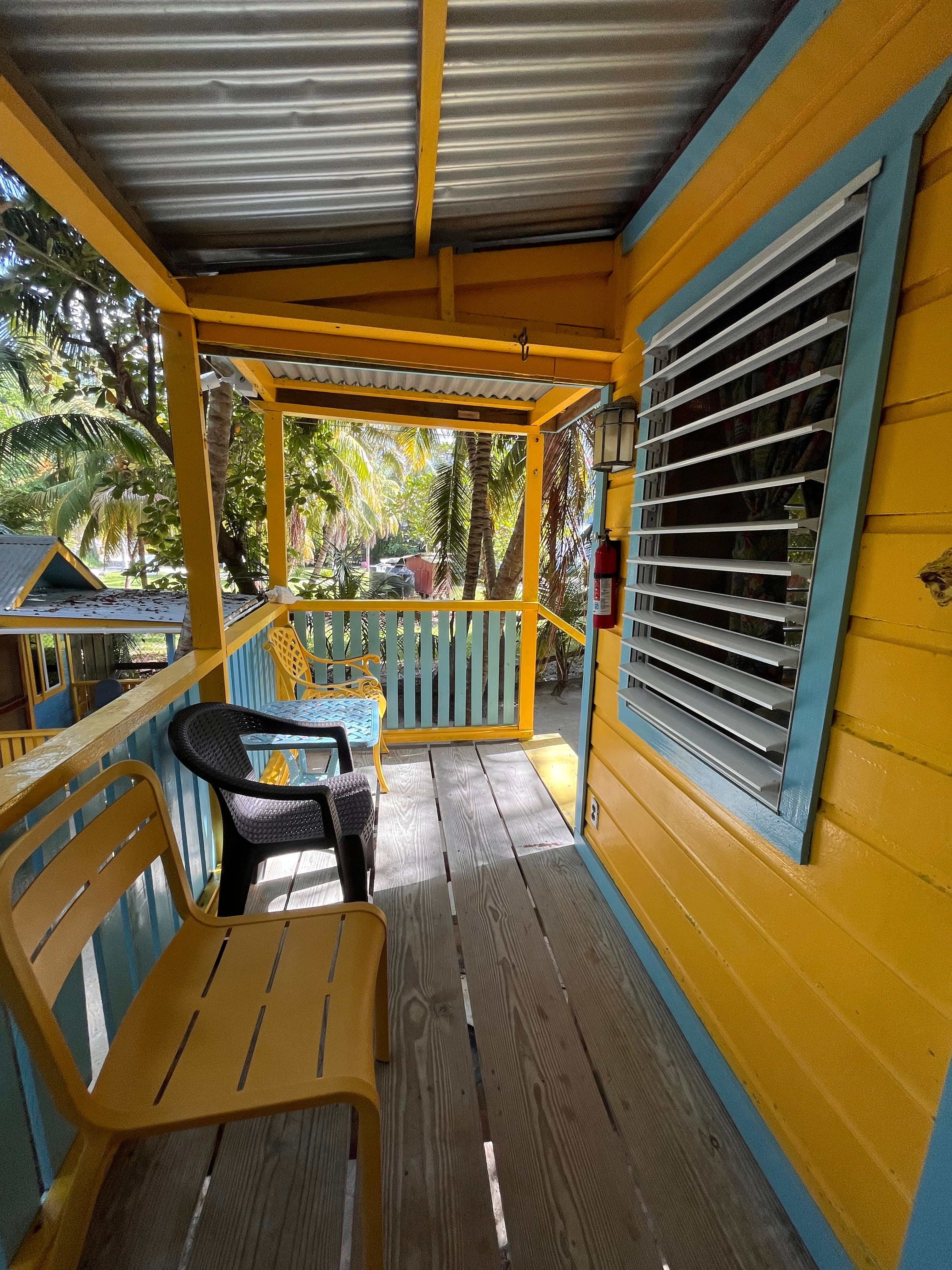 A brightly colored porch with yellow and blue accents, featuring two chairs and surrounded by tropical vegetation.