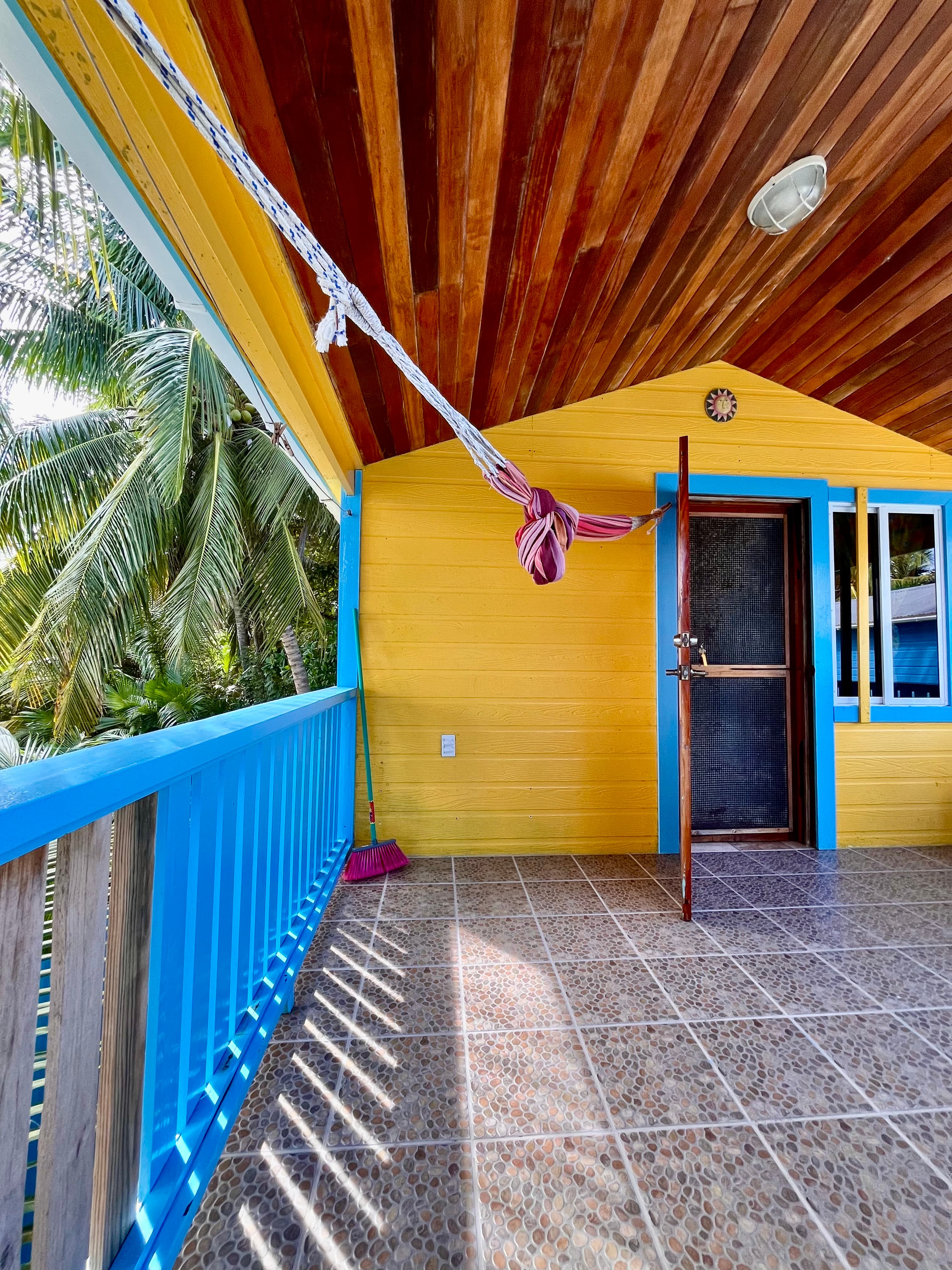 Brightly colored porch with a wooden ceiling, featuring a doorway and tropical plants in the background.