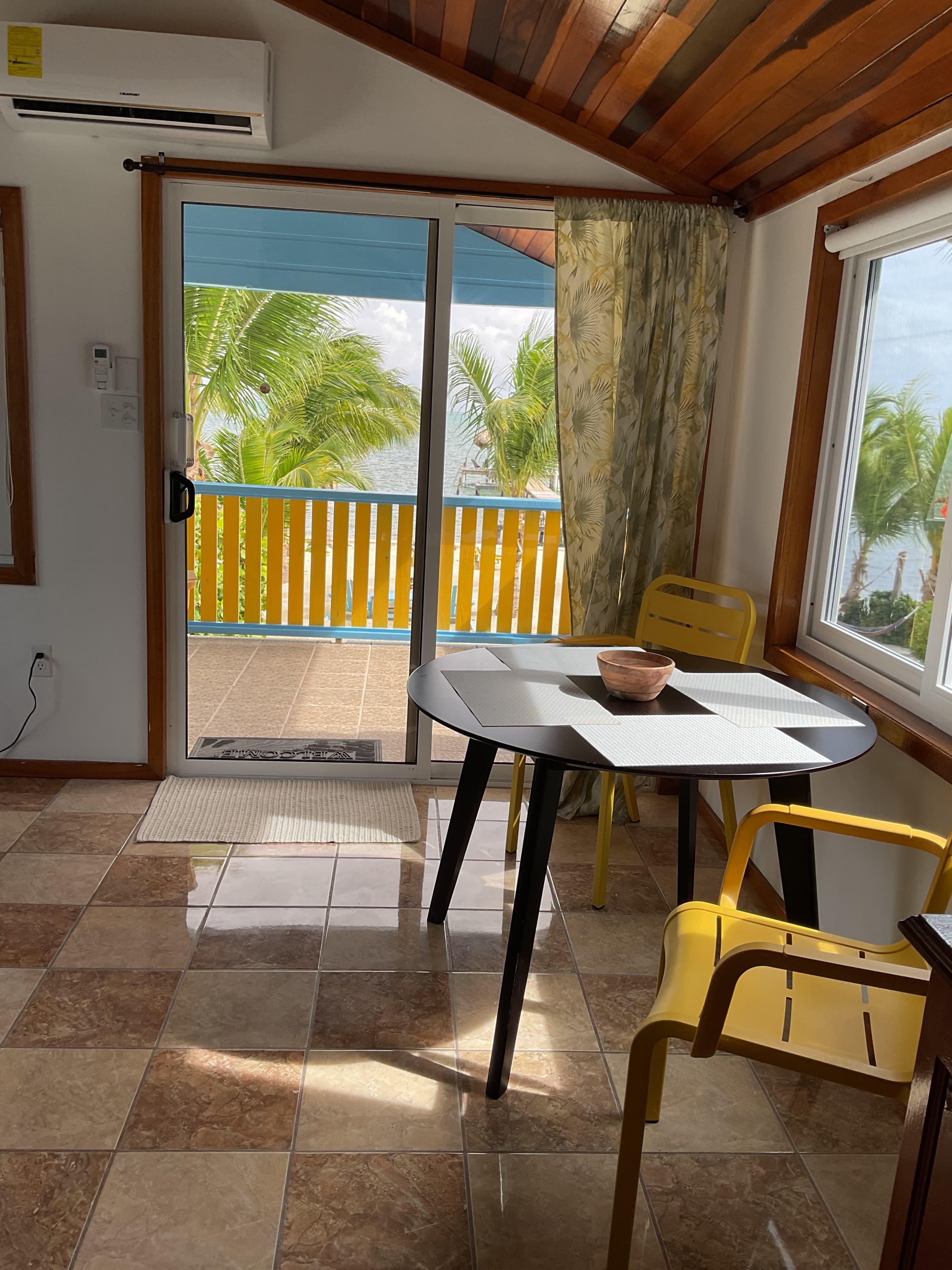 A sunlit room with a table and chairs, featuring a view of palm trees through a sliding glass door.