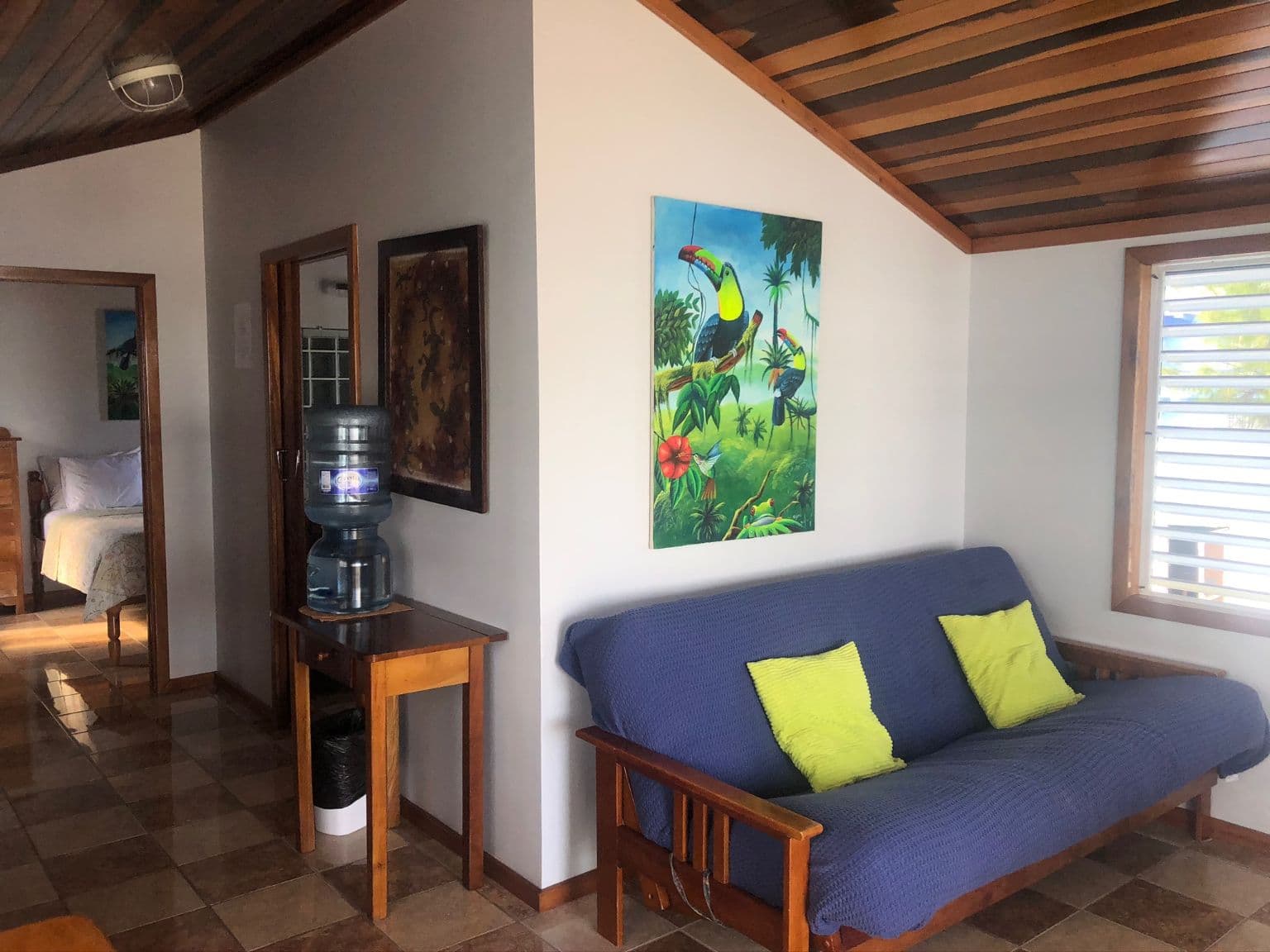 Sitting area with blue futon & yellow cushions, tropical-themed artwork, small table and water cooler nearby