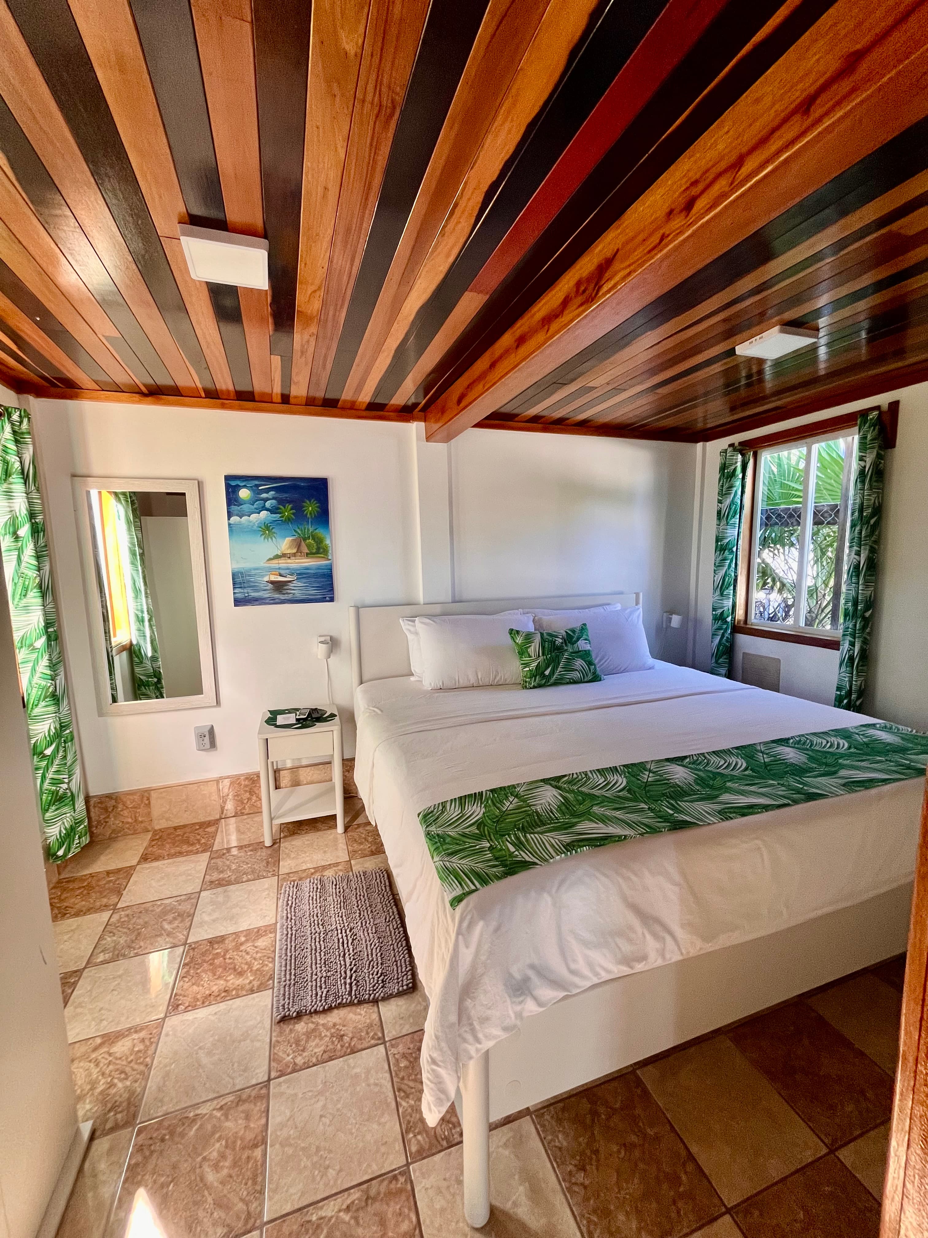 A cozy bedroom featuring a wooden ceiling, a double bed with leaf-patterned decor, and bright, tropical-themed curtains.