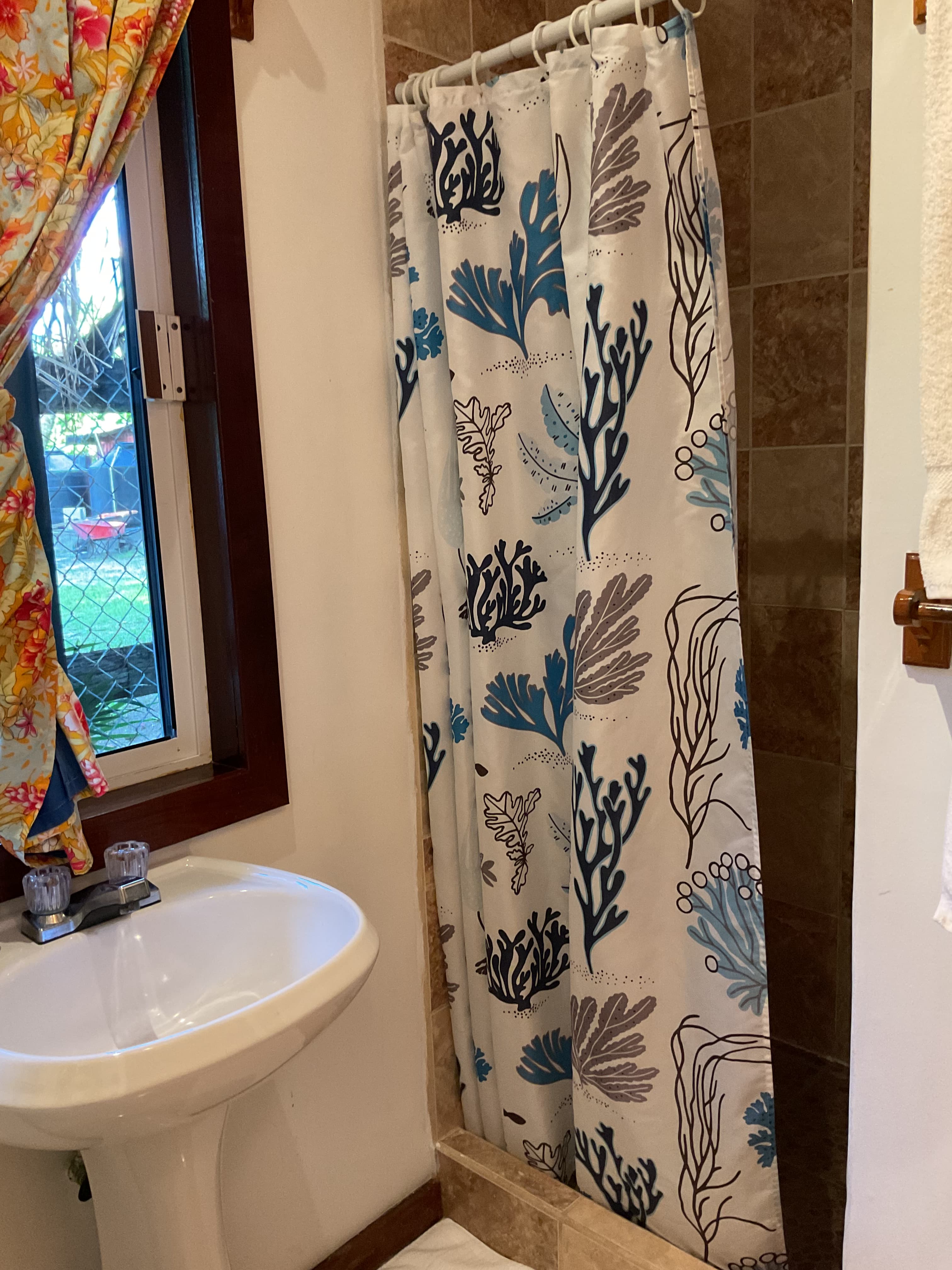 A bathroom with a decorative shower curtain, a white pedestal sink, and a window with floral curtains.