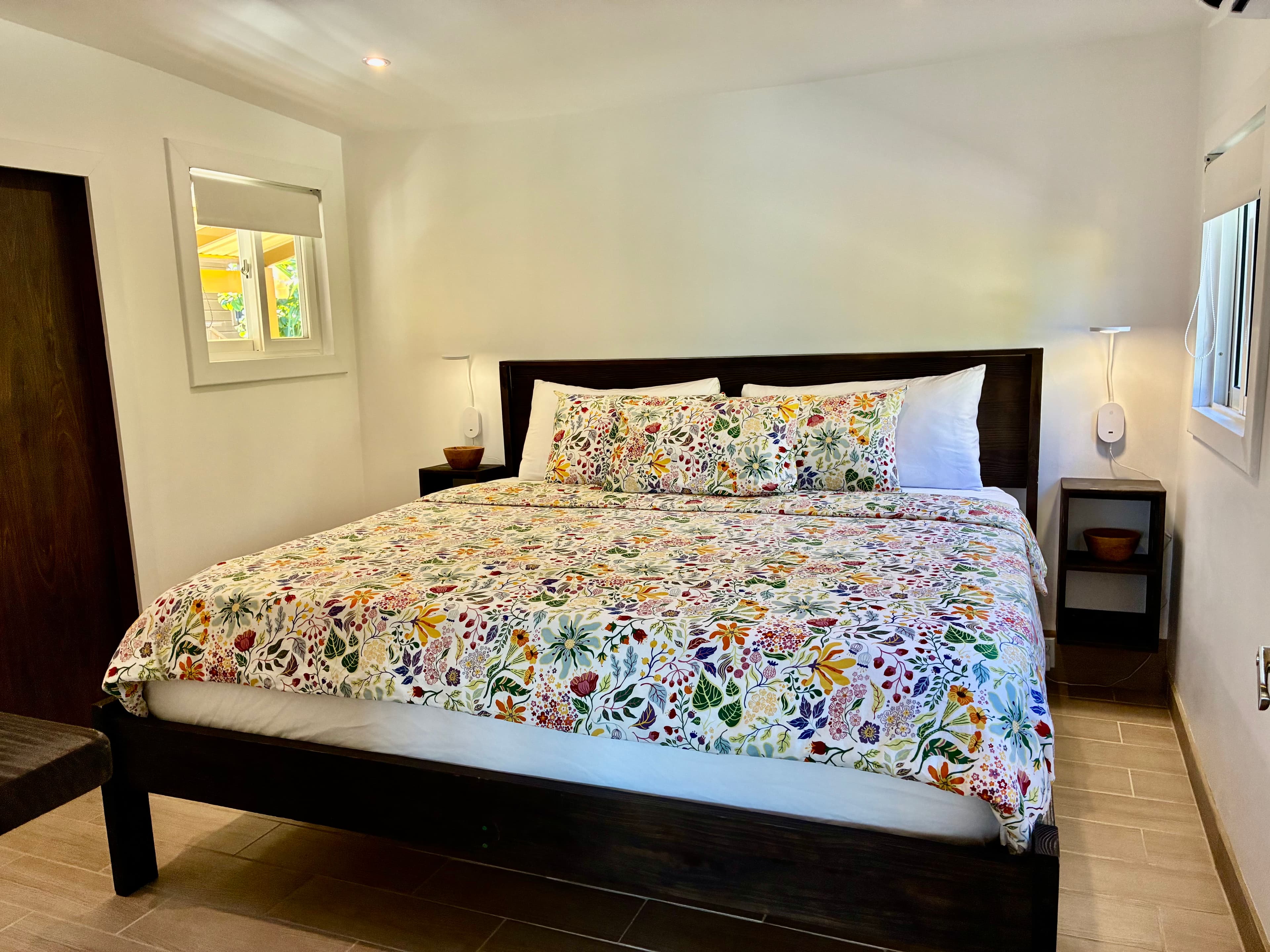 A brightly patterned floral bedspread covers a modern wooden bed in a cozy, well-lit room.