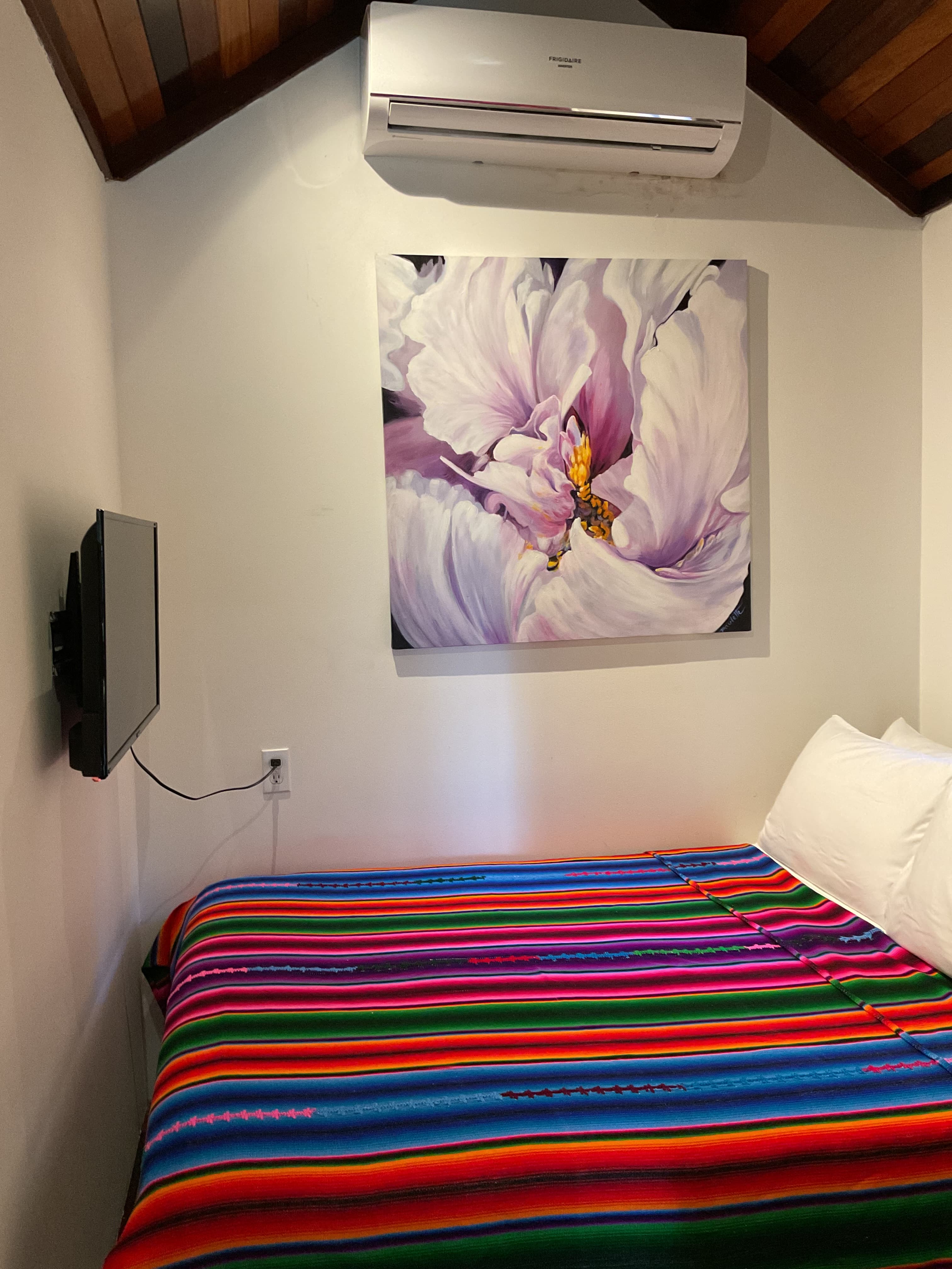 A colorful striped blanket covers a bed in a room with a wall-mounted TV and a large flower painting above it.