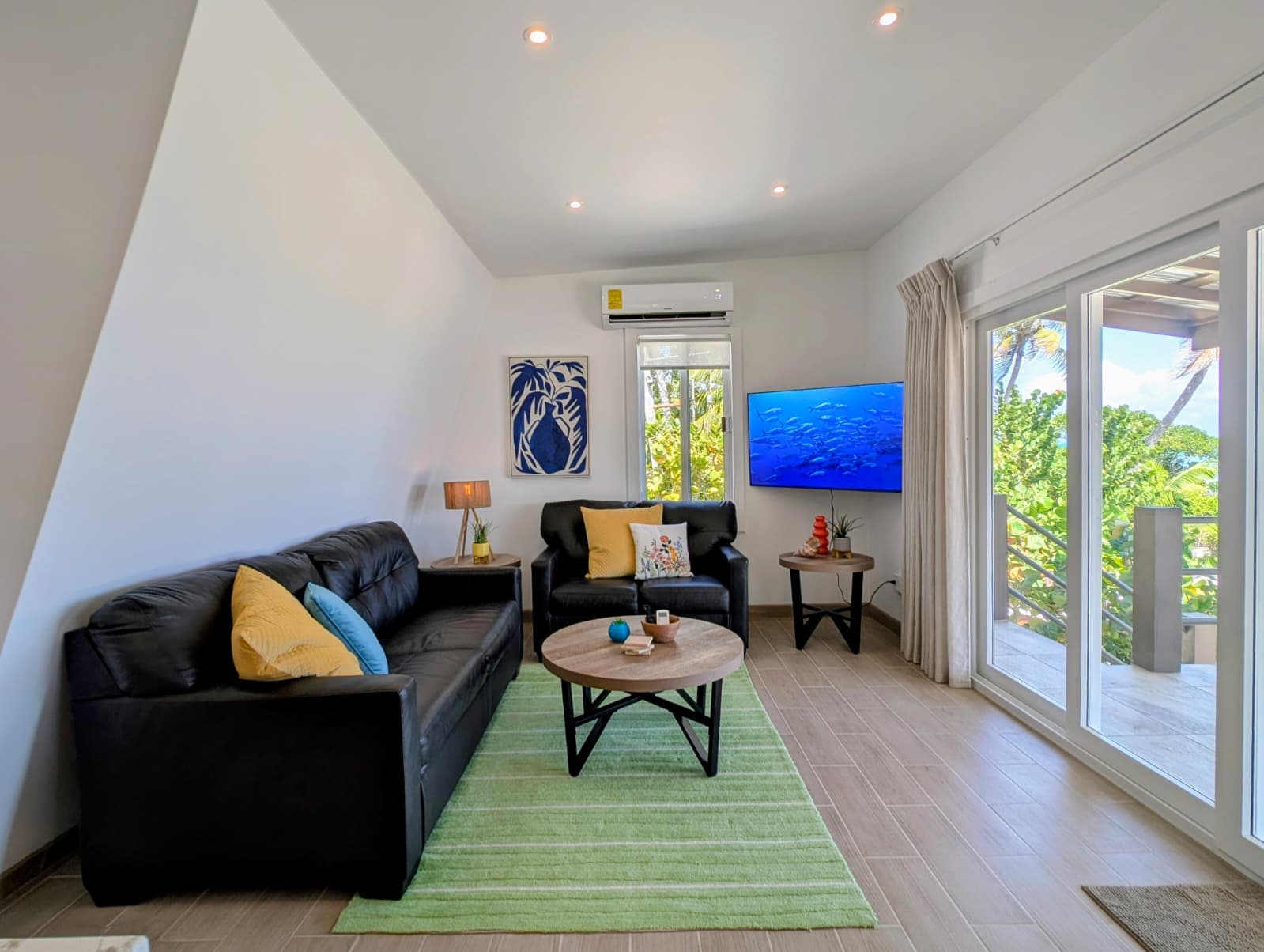 A bright and airy living room featuring a black sofa, wooden coffee table, and large windows opening to an outdoor view.